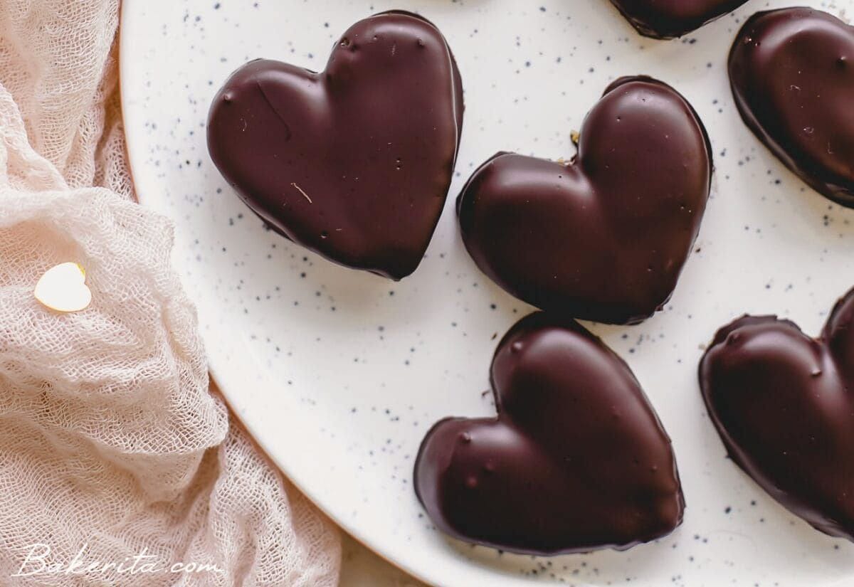 Dark chocolate–coated peanut butter hearts displayed on a speckled white plate, with soft fabric draped beside them.