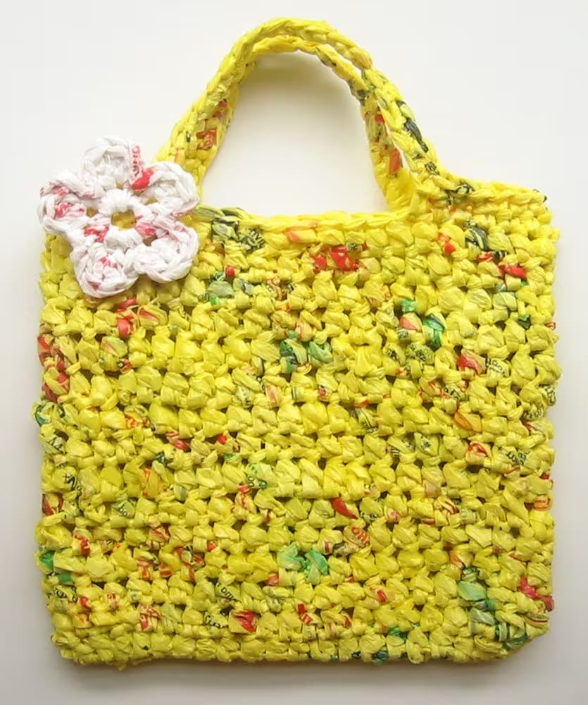 A small bag made from plastic yarn. The body is made from yellow plastic bags and it has a white plarn flower.
