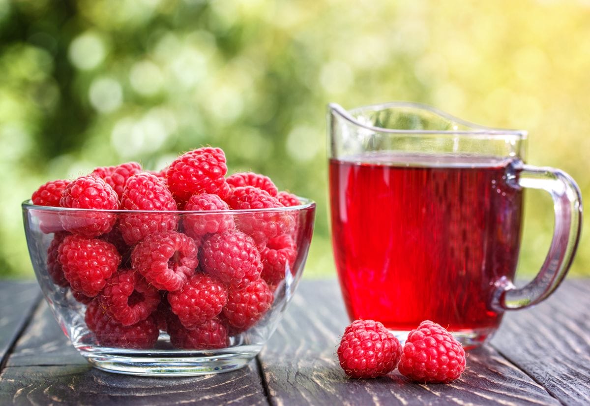 A pitcher of raspberry cordial and a bowl of fresh raspberries on a wooden table outdoors.
