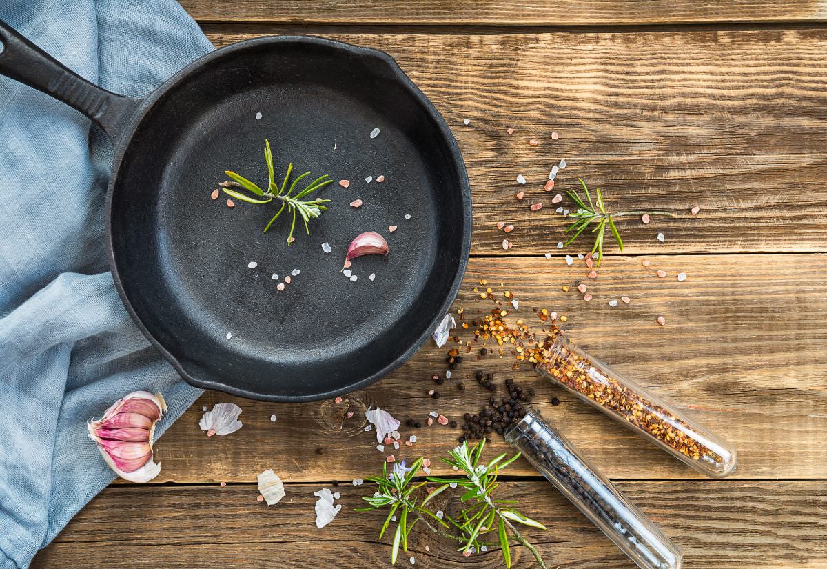 A cast iron skillet surrounded by fresh herbs, garlic, and spices on a rustic wooden table.