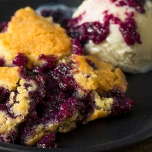 A close up view of warm blueberry cobbler and a scoop of vanilla ice cream on a black plate.