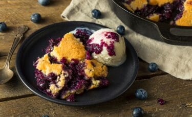 Warm blueberry cobbler with vanilla ice cream served on a black plate.