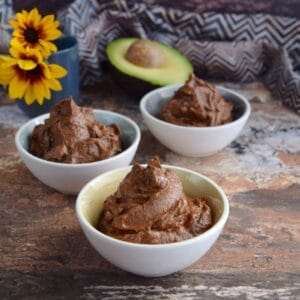 Three small bowls filled with creamy avocado chocolate pudding on a rustic table, with a halved avocado and decorative sunflowers in the background.