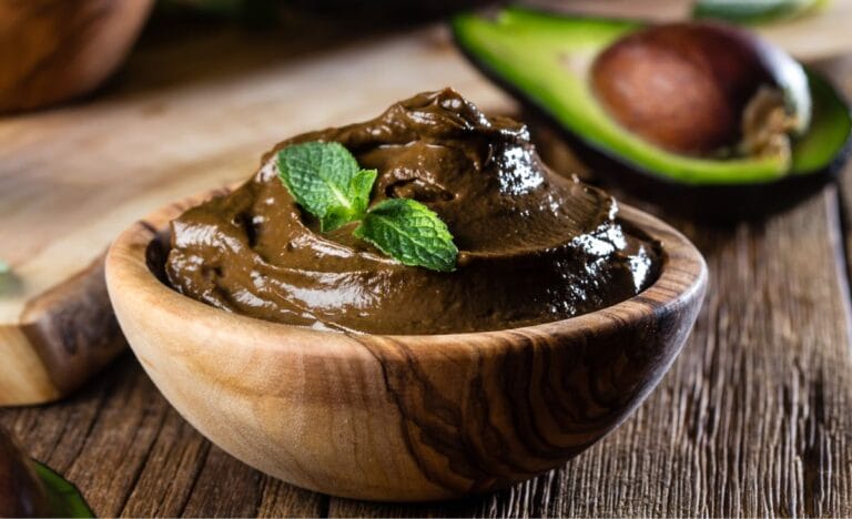 A creamy bowl of avocado chocolate pudding garnished with fresh mint leaves, with an avocado in the background.