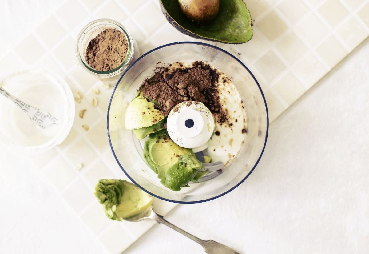 Ingredients for avocado chocolate pudding, including avocados, cocoa powder, and milk, in a food processor.