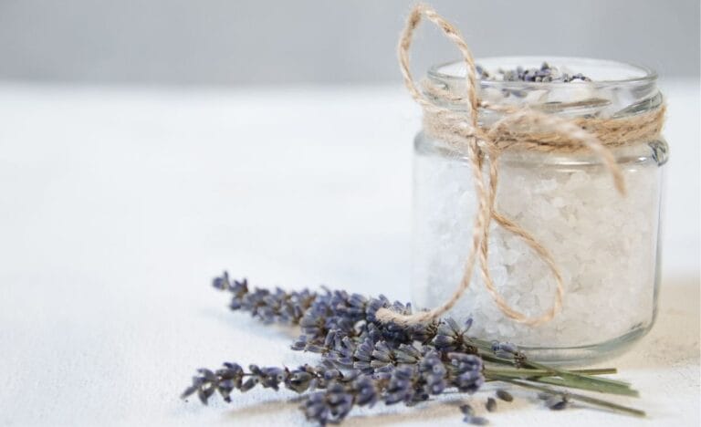 A glass jar filled with sea salt bath soak, tied with rustic twine, accompanied by dried lavender sprigs on a white background.