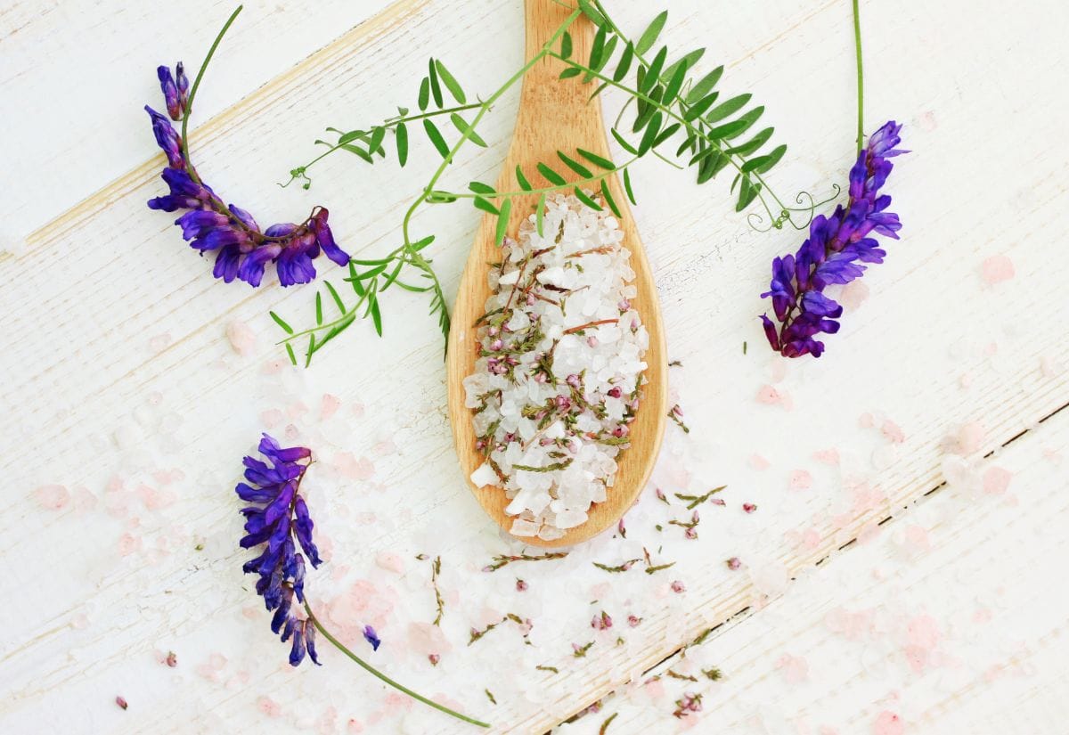 A wooden spoon holding a mix of bath salts and dried herbs, surrounded by delicate purple flowers on a whitewashed wooden surface.