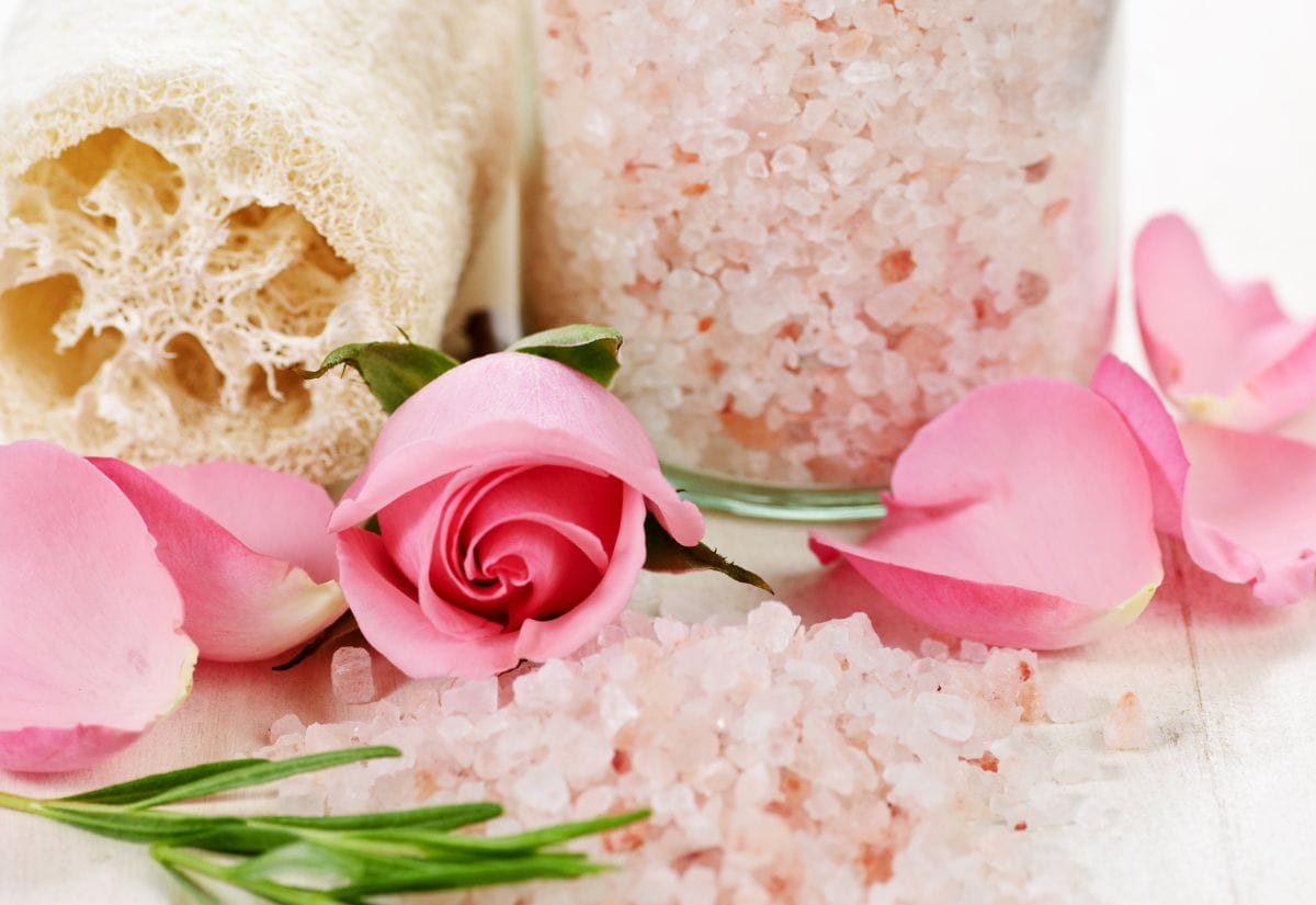 A jar of pink Himalayan bath salts with fresh rose petals, rosemary sprigs, and a loofah on a clean white background.