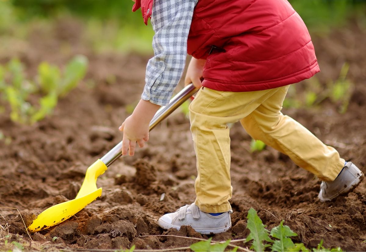 A young child digging in a garden bed with a yellow toy shovel.