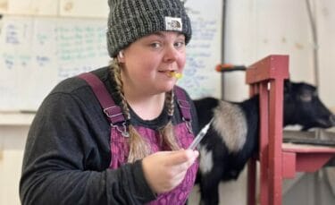 Carrissa from Lone Feather Farm wearing a beanie and overalls, holding a syringe with a playful smile, standing next to a goat.