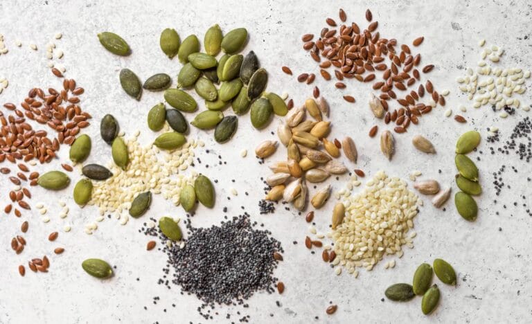 A variety of vegetable seeds, including pumpkin, flax, and sesame, scattered on a light surface.