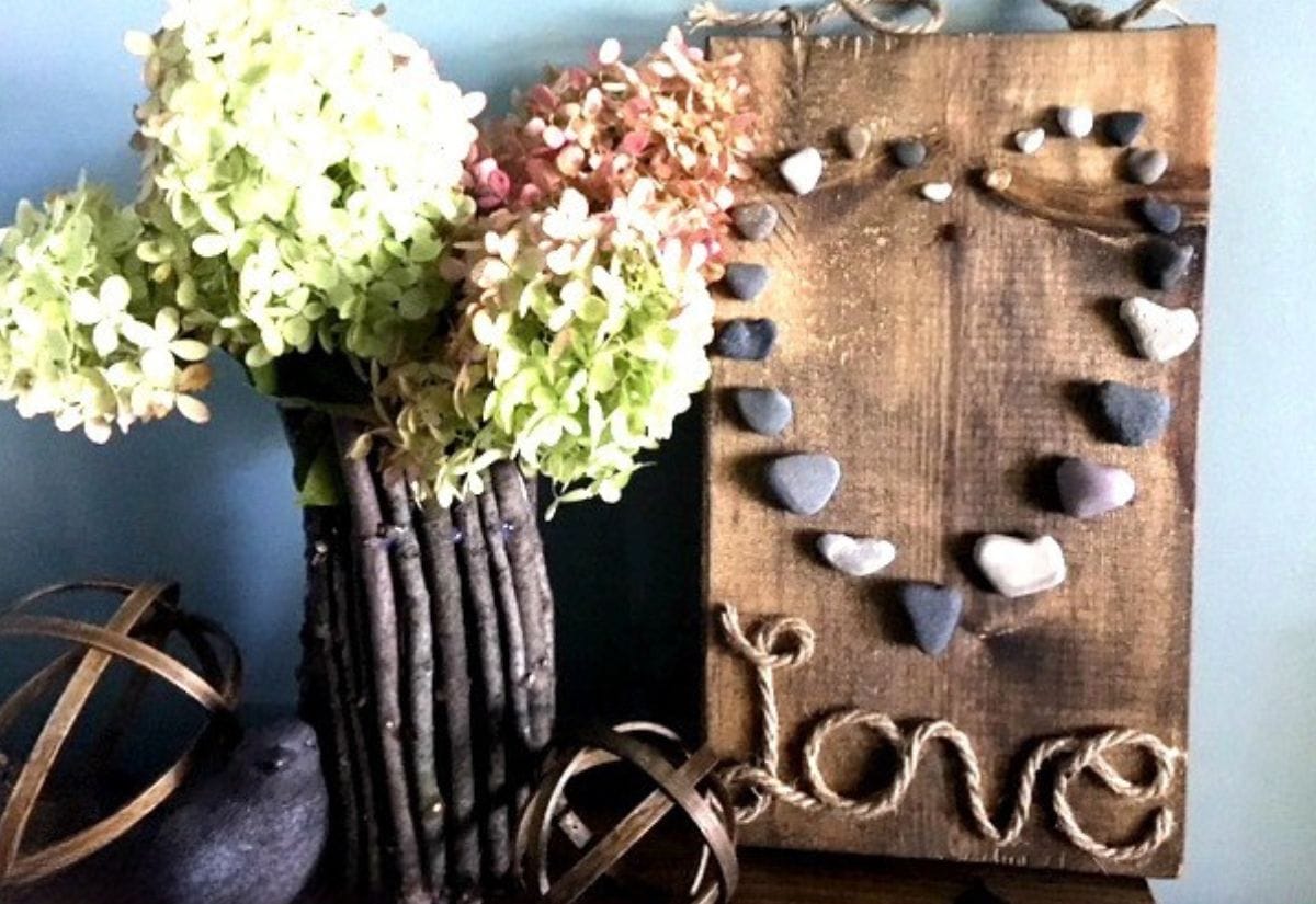 A wooden sign adorned with a heart-shaped pebble design and twine spelling out "Love," placed next to a bouquet of dried hydrangeas.