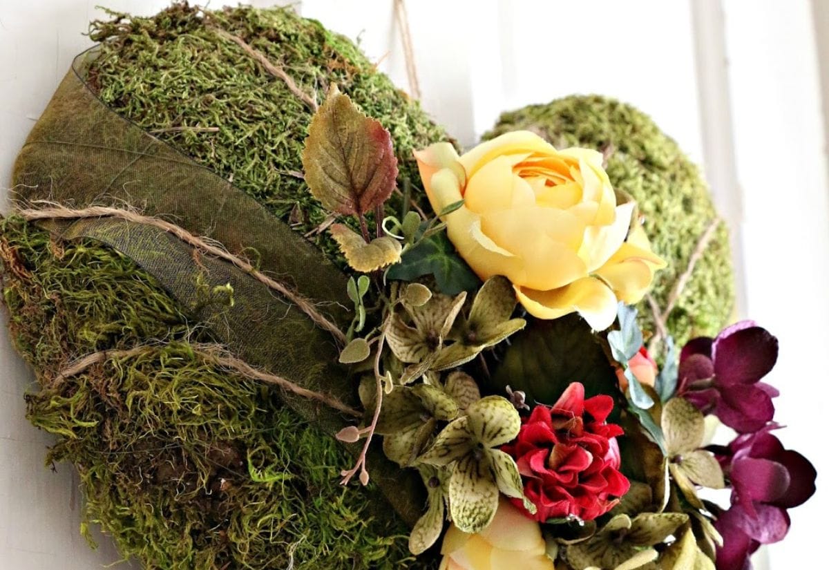 A moss-covered heart wreath adorned with faux yellow roses and red flowers, tied with rustic twine.