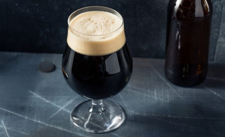 A glass of freshly brewed Irish stout with a creamy tan head, sitting on a dark rustic surface next to a brown bottle.