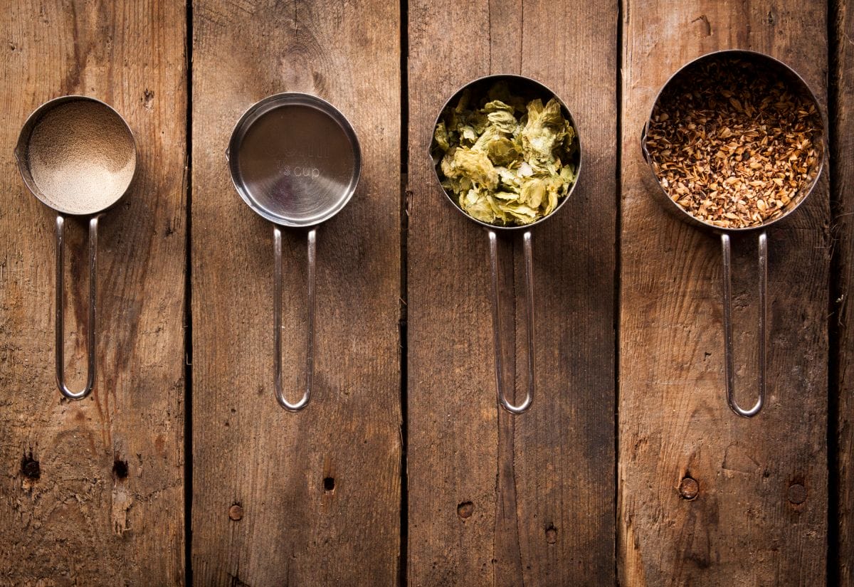 Measuring cups filled with brewing ingredients: yeast, hops, malted barley, and specialty grains.