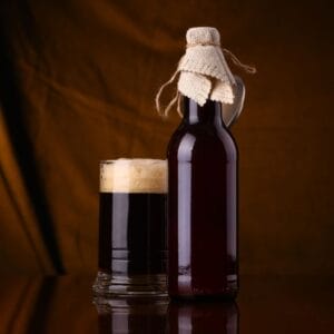 A glass of dark Irish stout with a frothy head next to a brown bottle wrapped with rustic burlap, set against a warm brown background.