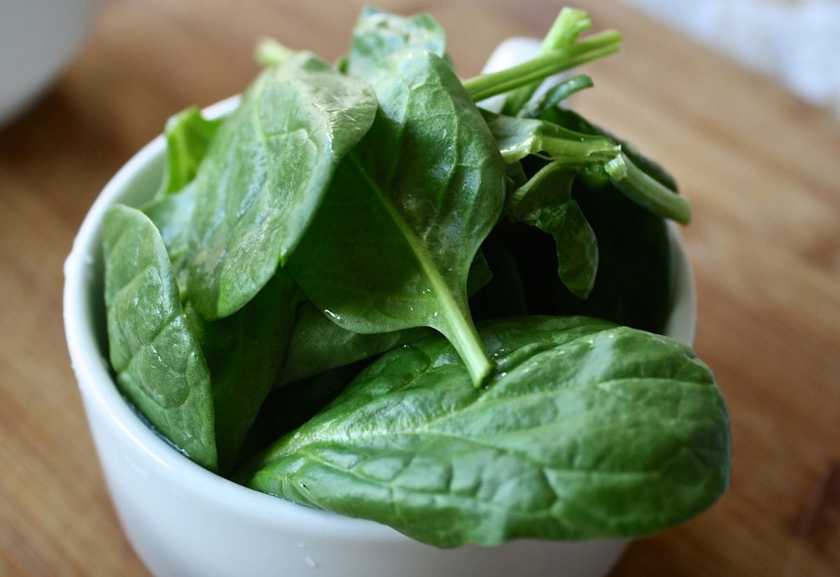 A small white bowl filled with fresh spinach leaves on a wooden cutting board, ready for blending into homemade green food coloring.