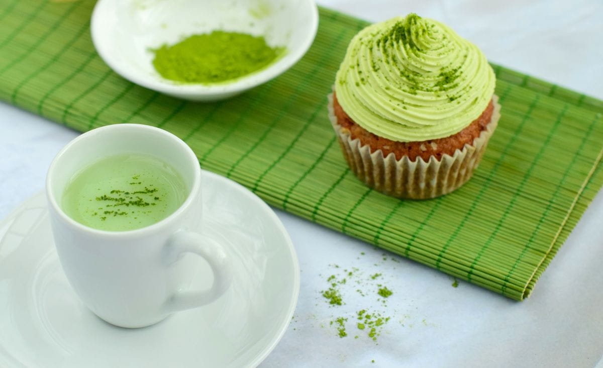 A matcha cupcake with green frosting and a cup of matcha latte, both dusted with matcha powder on a green bamboo mat.