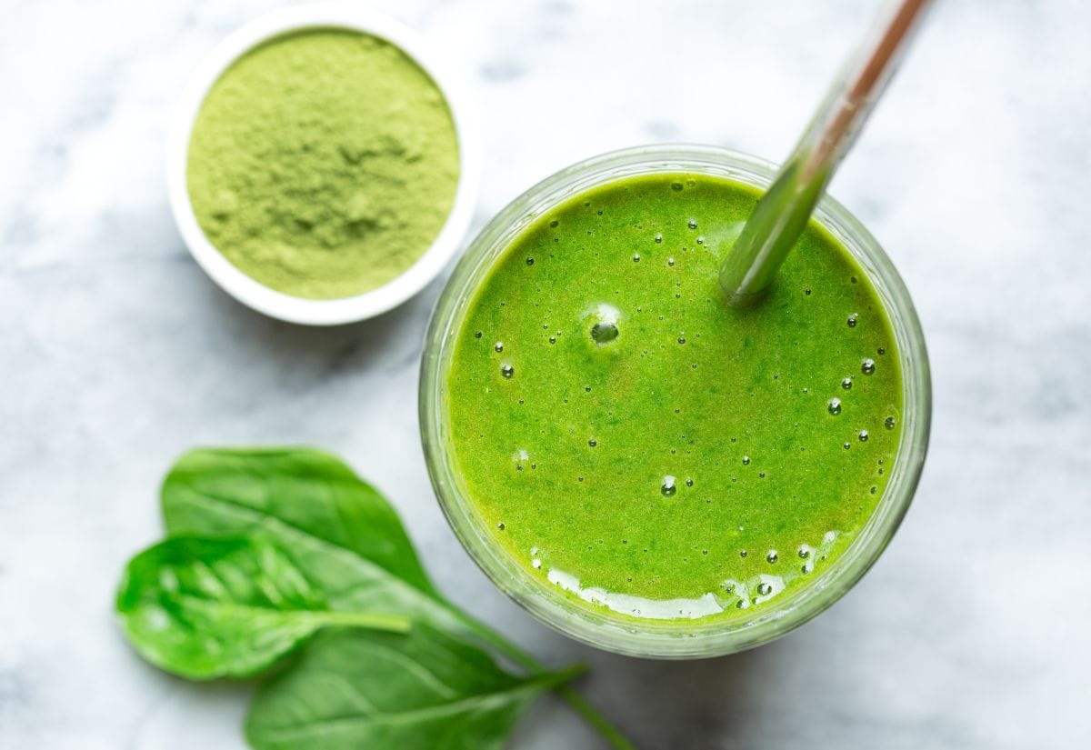 A vibrant green smoothie in a glass with a bamboo straw, surrounded by fresh spinach leaves and a bowl of matcha powder.