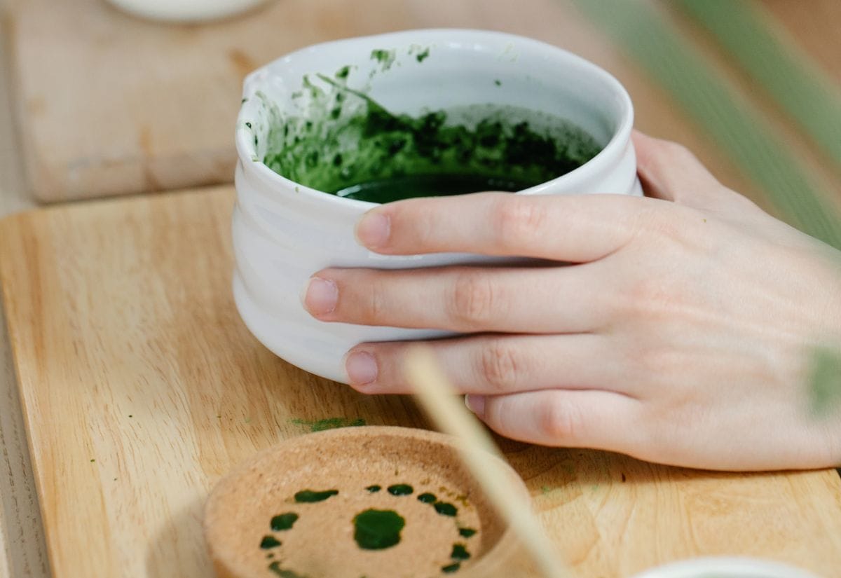 A hand holding a white bowl of fresh homemade green food coloring made from matcha, with a wooden spoon resting nearby.