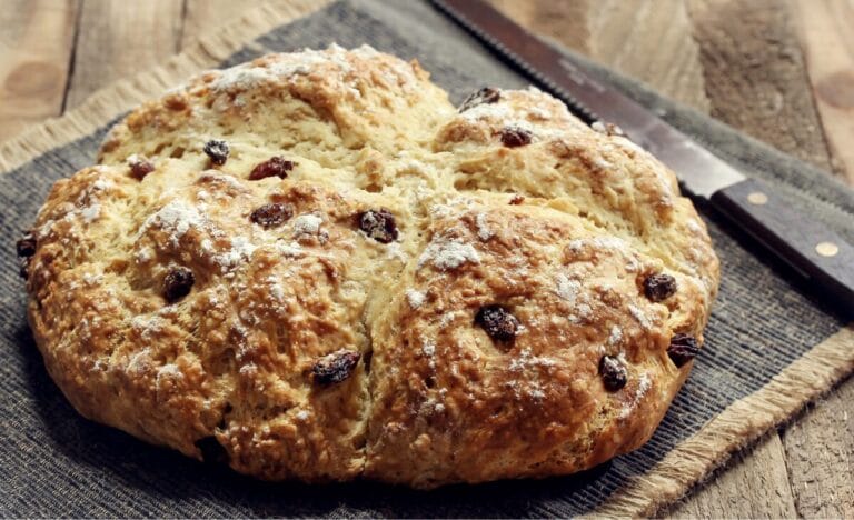 A freshly baked Irish soda bread with a golden-brown crust and raisins, sitting on a rustic wooden surface with a serrated knife nearby.