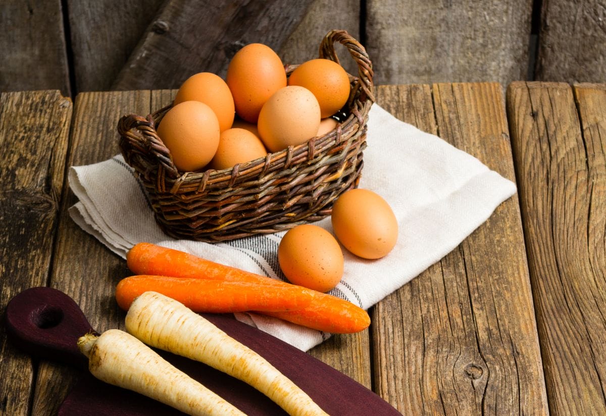 A rustic basket of fresh eggs alongside carrots and parsnips on a wooden table.