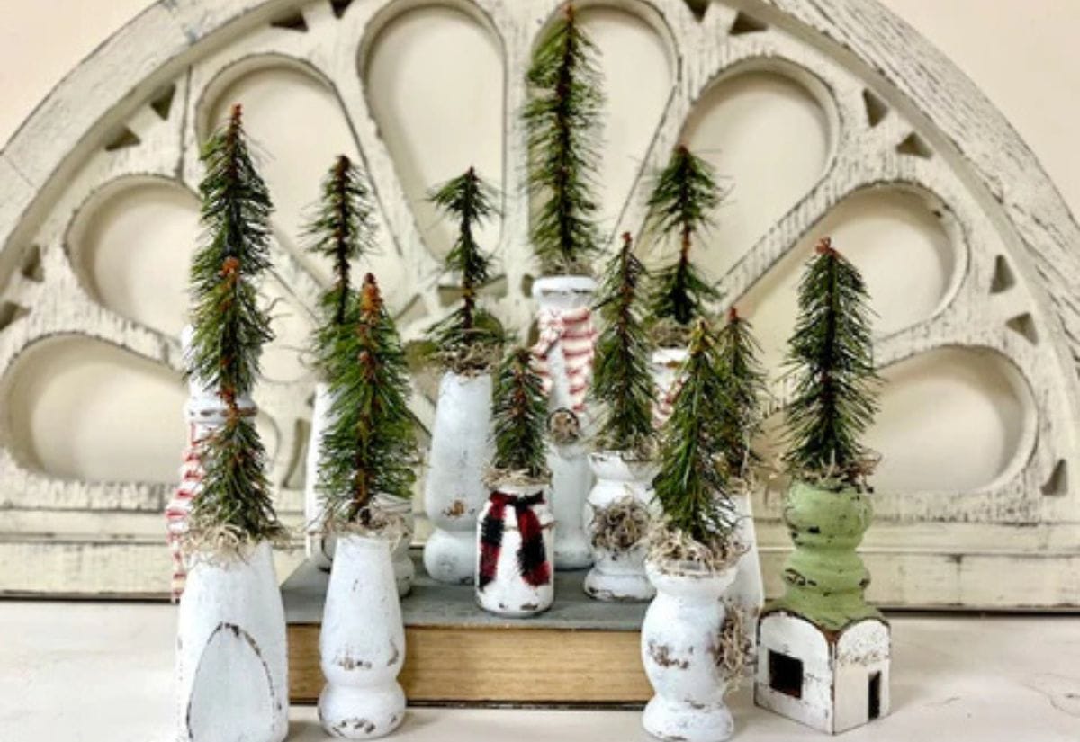 A charming collection of upcycled wooden spindles painted in distressed white, each topped with a small faux pine tree, arranged in a rustic winter village display.