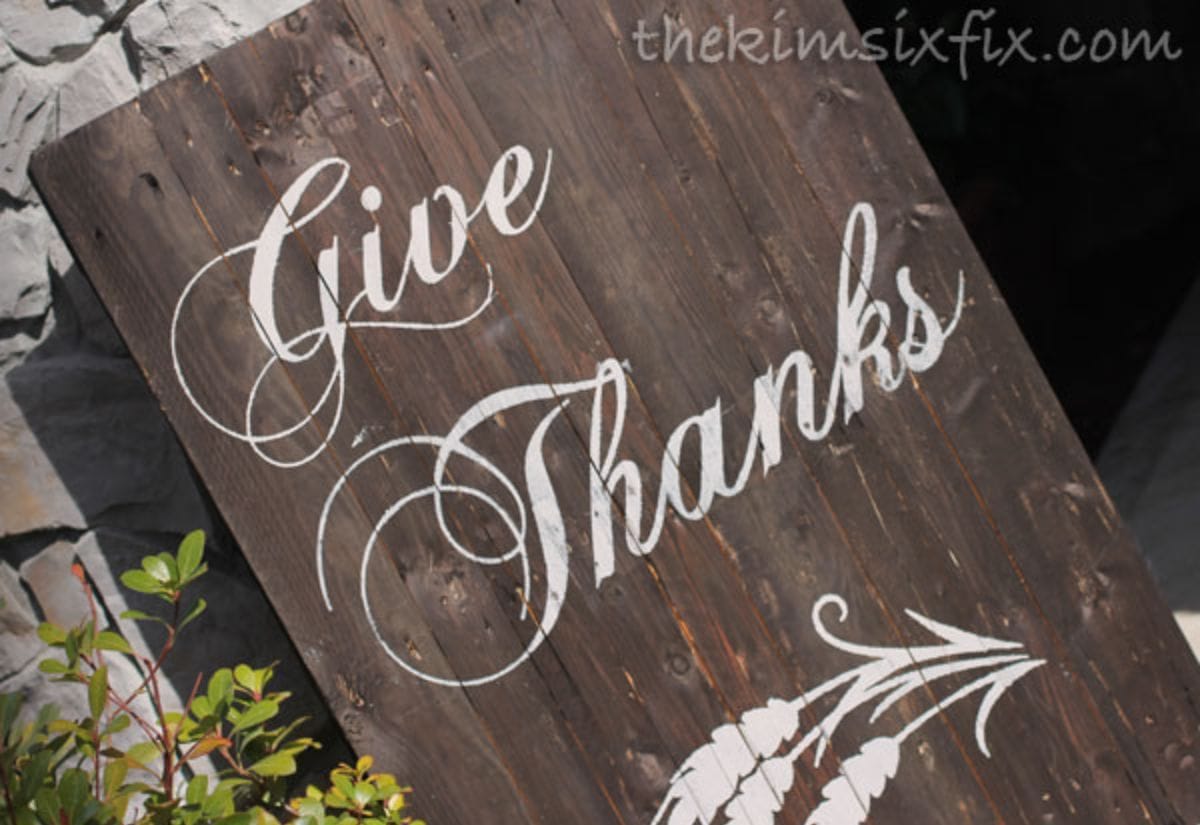 A reclaimed wood sign with "Give Thanks" hand-painted in elegant script, leaning against an outdoor stone wall for a charming farmhouse look.