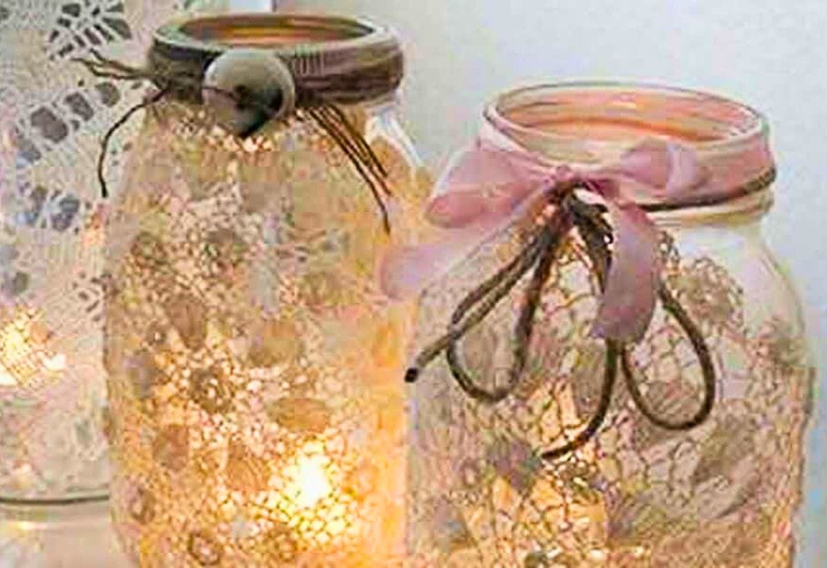 Two upcycled mason jars wrapped in delicate lace, adorned with twine and ribbon, glowing warmly from candlelight inside.