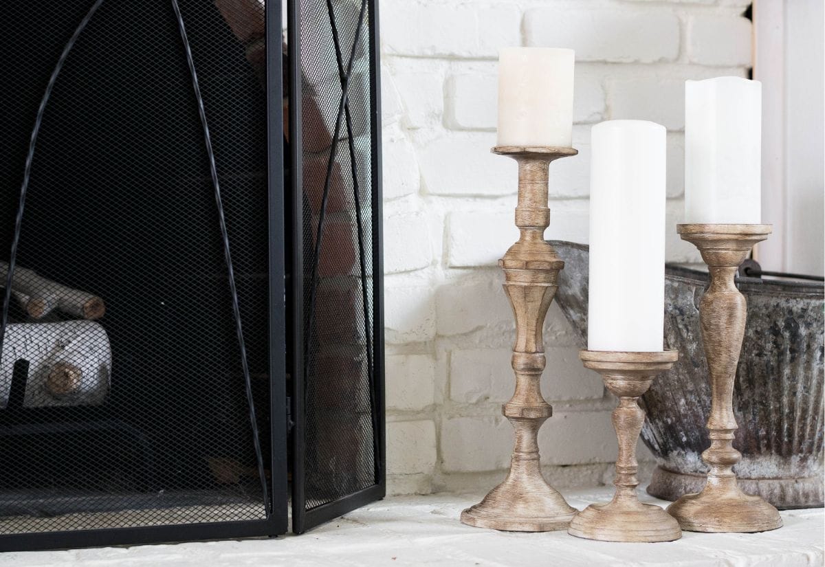 Three wooden candlesticks of varying heights with a weathered farmhouse finish, placed beside a fireplace with white pillar candles.