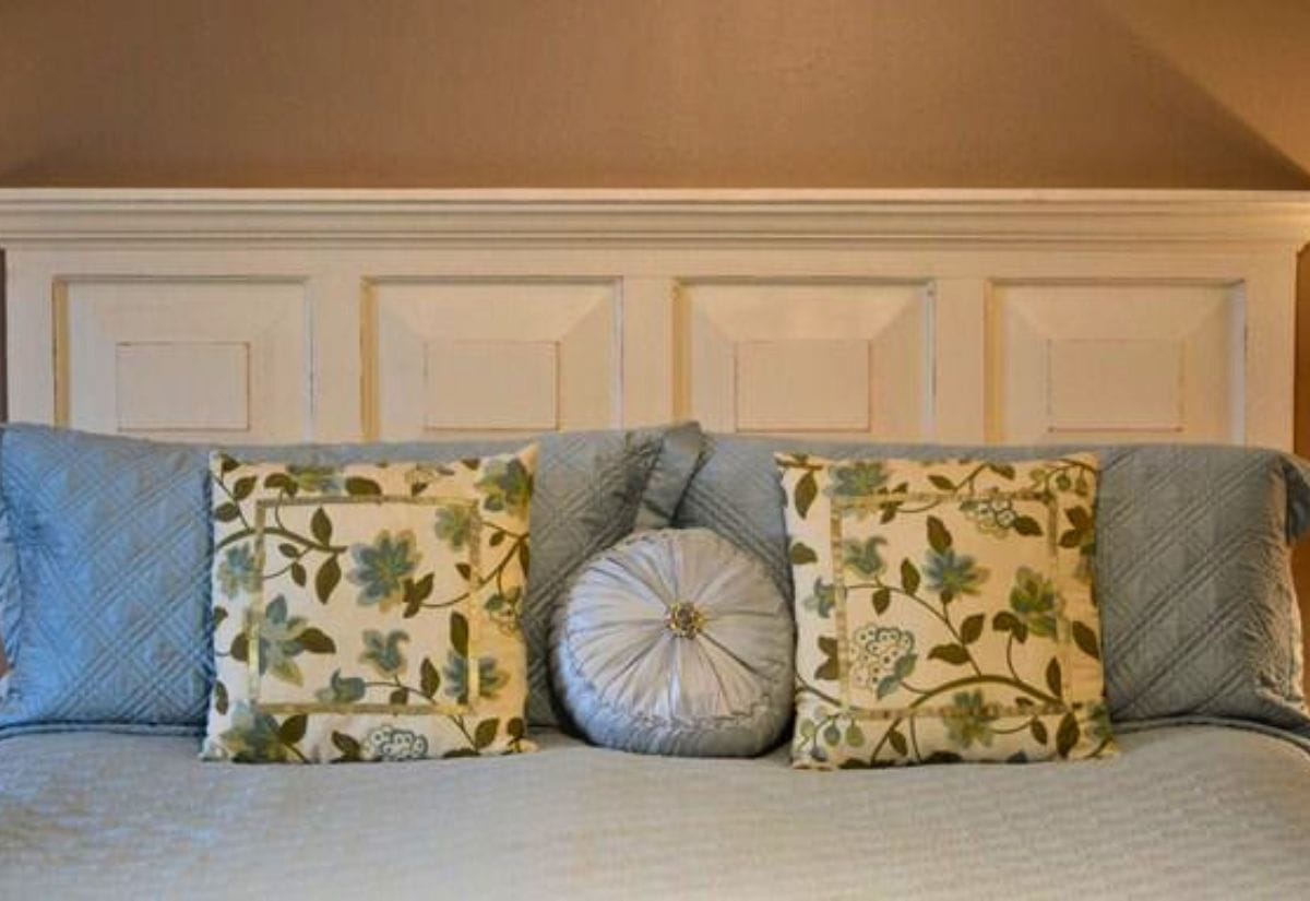 A cozy bedroom featuring a headboard made from a repurposed old door, painted in soft white, with floral and solid blue pillows.