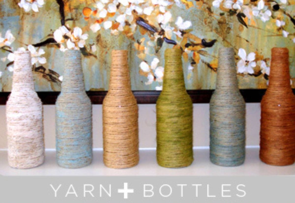 A row of upcycled glass bottles wrapped in neutral and pastel-colored yarn, creating rustic farmhouse vases.