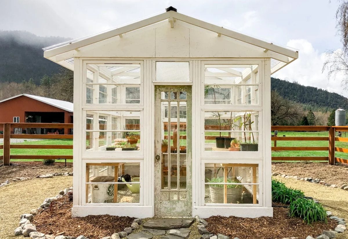 A charming greenhouse built entirely from repurposed old windows and doors, painted white, standing in a picturesque farm setting.