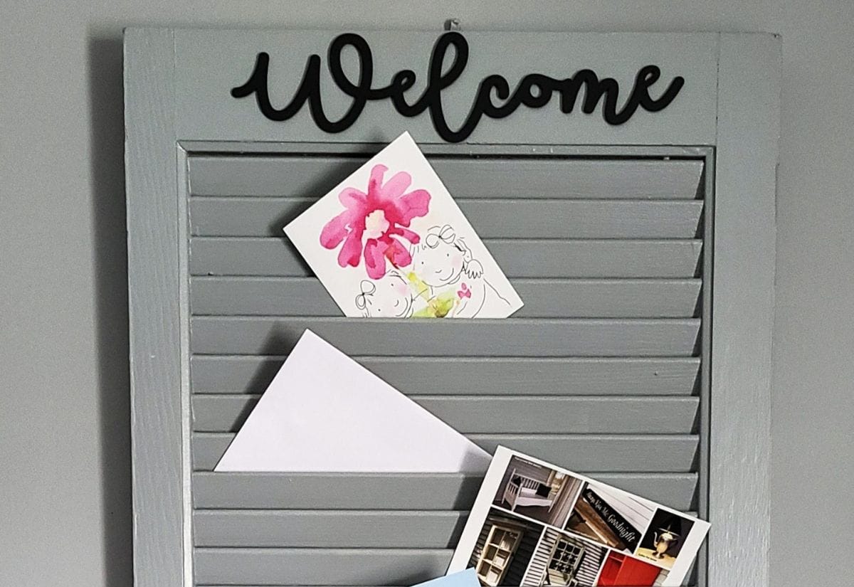 A gray wooden shutter transformed into a farmhouse-style mail and note organizer, decorated with a black "Welcome" sign and clipped postcards.