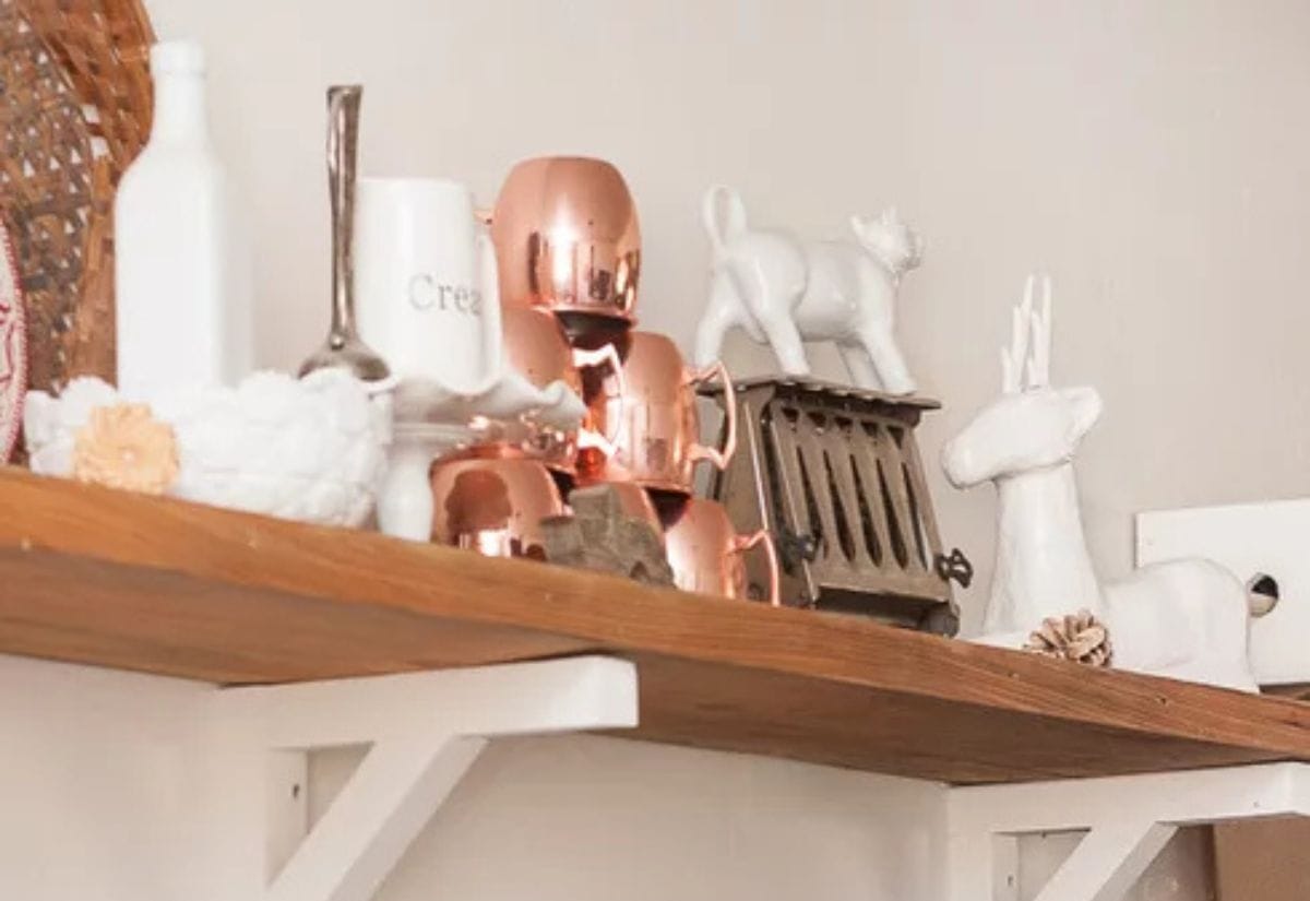 A rustic open shelf displaying an assortment of upcycled decor, including white ceramic figurines, vintage copper mugs, and woven baskets.