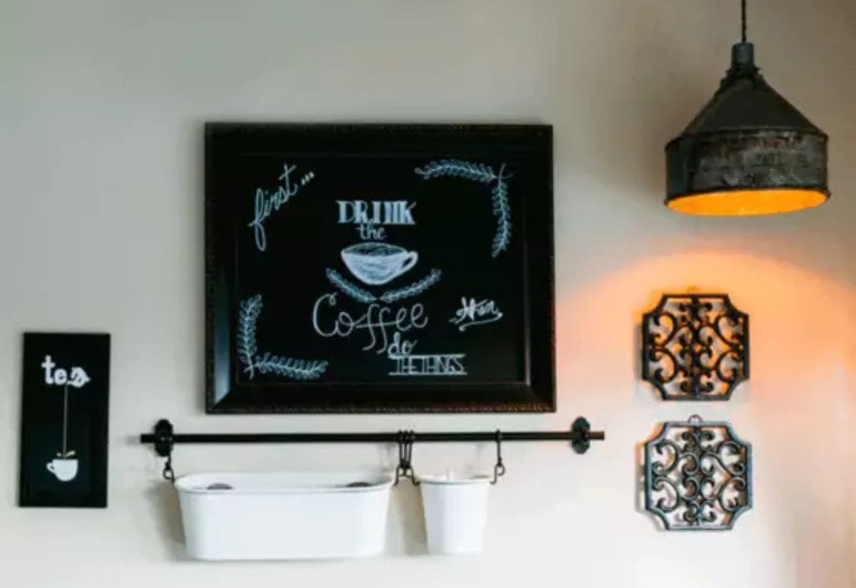 A cozy farmhouse kitchen wall display featuring a vintage chalkboard sign, ornate metal wall art, a hanging storage rail, and a rustic industrial light fixture.