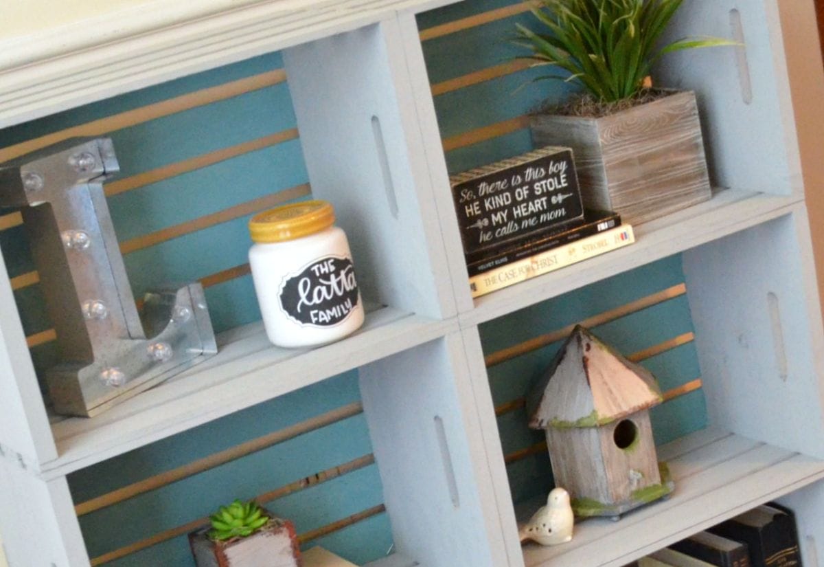 A repurposed wooden crate bookshelf painted in distressed blue, styled with rustic decor including books, plants, a birdhouse, and a personalized jar.