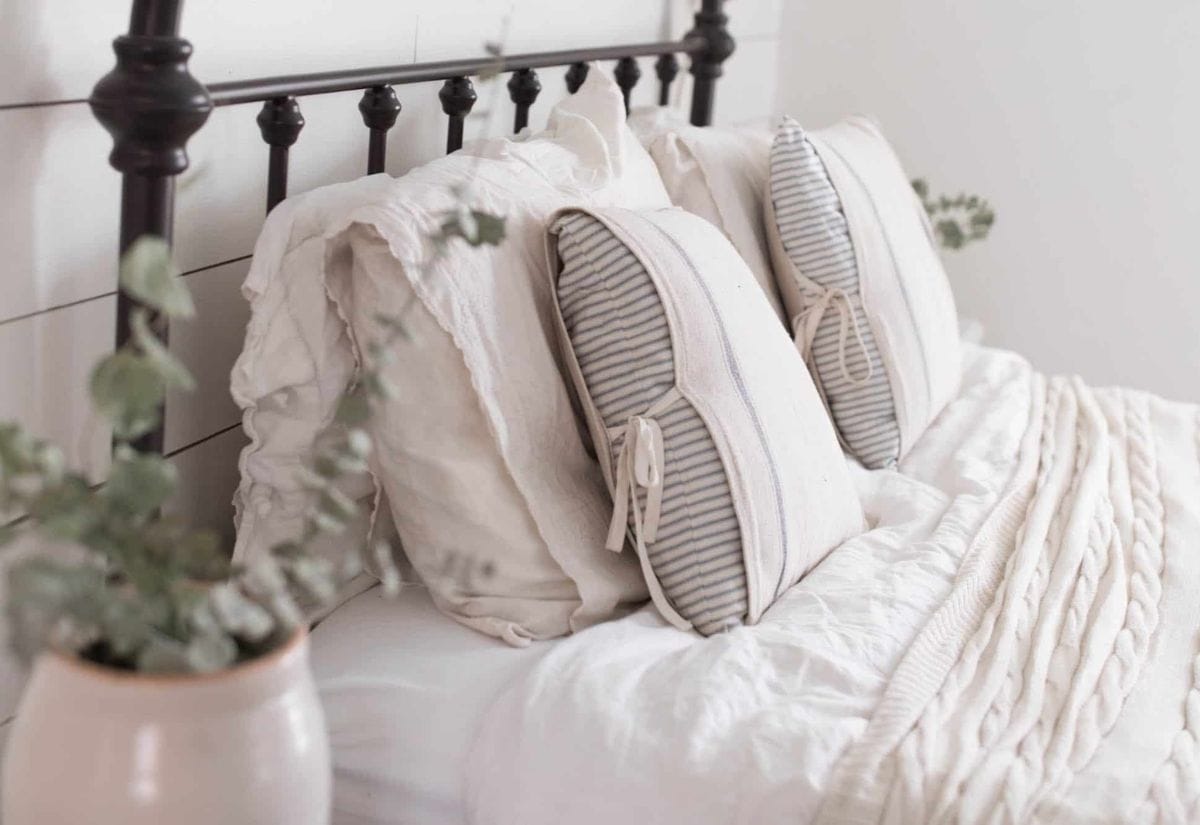 A beautifully styled farmhouse bedroom with an iron bedframe, white ruffled bedding, striped tie-on pillows, and a ceramic vase with eucalyptus.