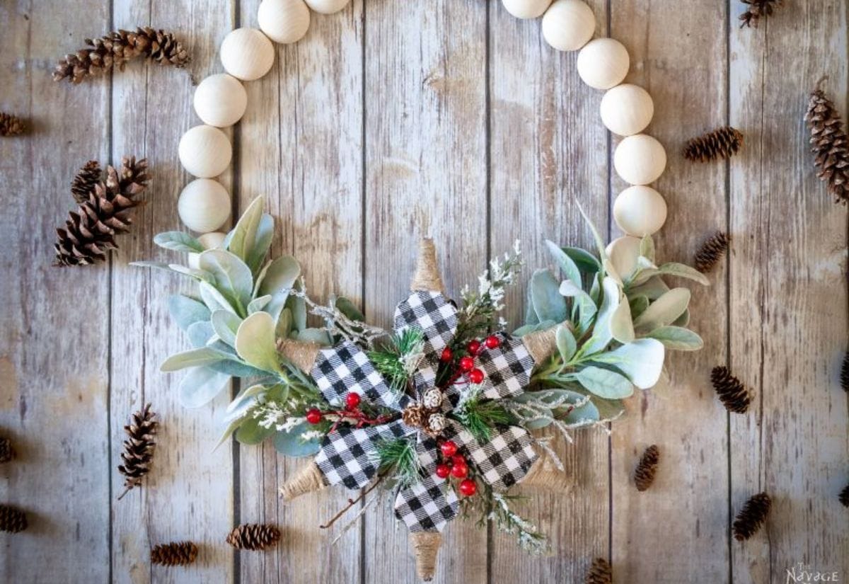 A handmade farmhouse-style wreath featuring wooden beads, greenery, and buffalo plaid ribbons, set on a rustic wood background with scattered pinecones.