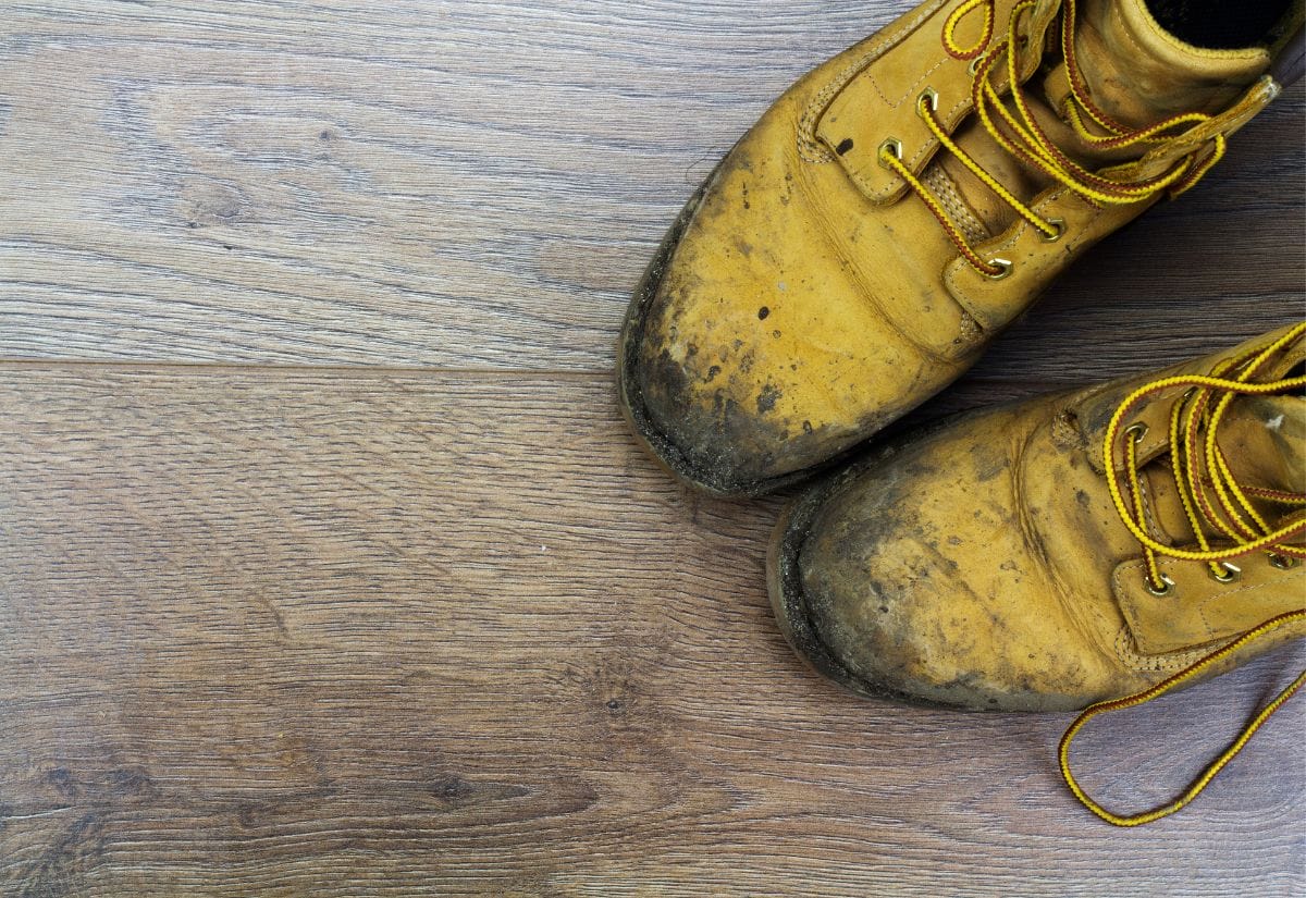 A pair of well-worn yellow work boots covered in dried mud sitting on a wooden floor, a reminder of the daily cleanup routine in spring.