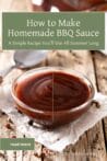 A small glass bowl filled with rich homemade BBQ sauce sitting on parchment paper on a rustic wood table, surrounded by peppercorns and a metal spoon.