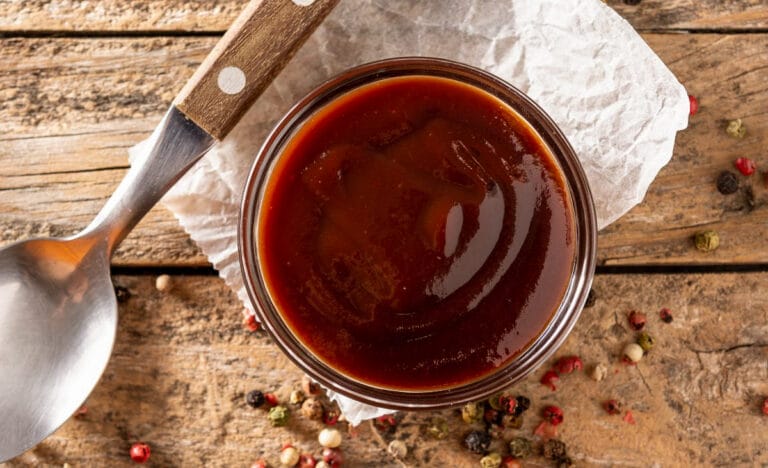 Overhead view of homemade BBQ sauce in a small glass bowl with a rustic spoon and peppercorns scattered on a wooden table.