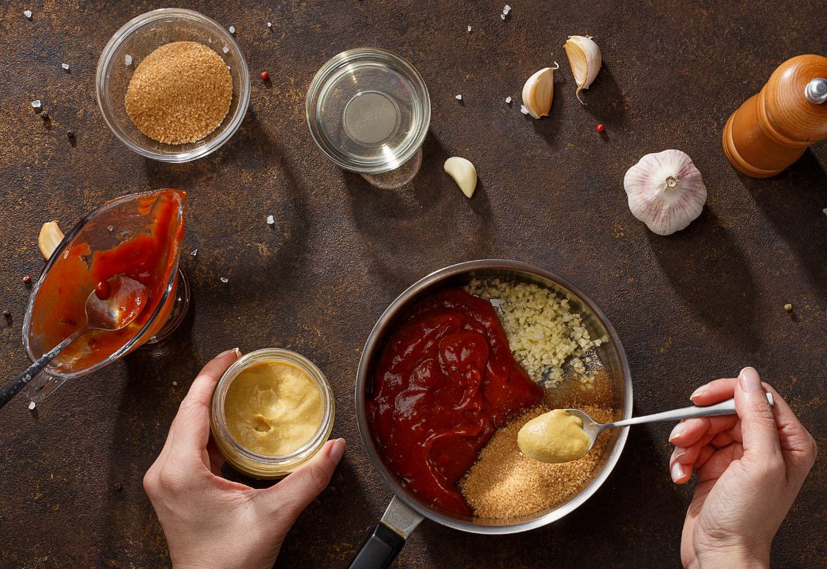 Ingredients for homemade BBQ sauce including ketchup, mustard, brown sugar, and minced fresh garlic arranged around a small saucepan.