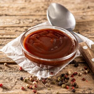Side view of a glass bowl filled with glossy homemade BBQ sauce, surrounded by colorful peppercorns on rustic parchment.