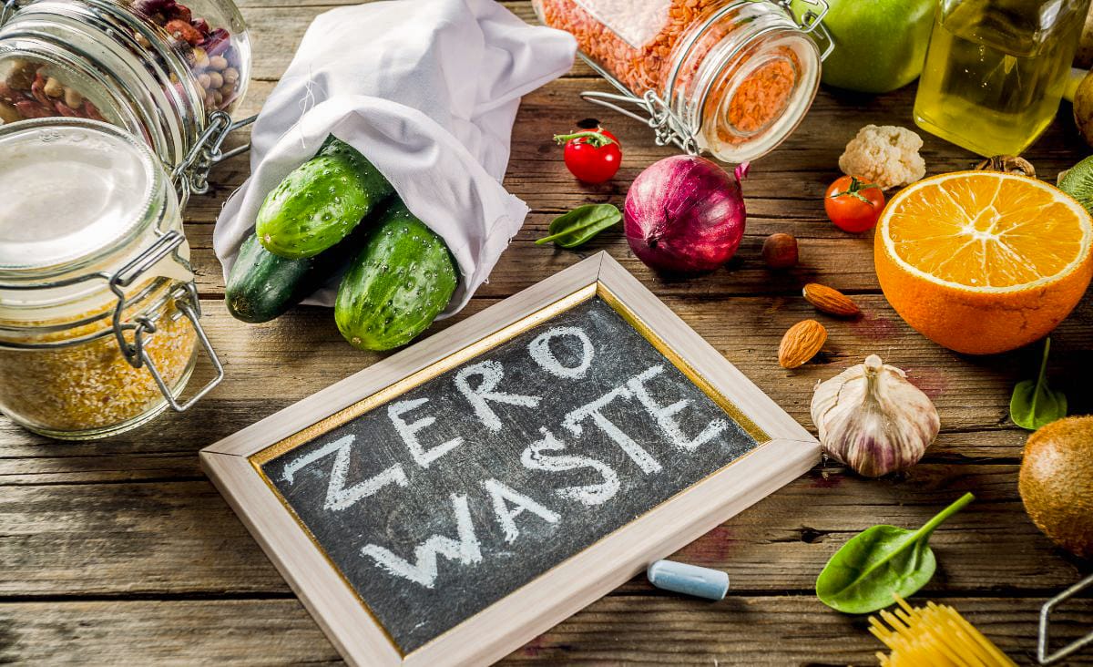 Overhead view of zero waste pantry staples including cucumbers in a cloth bag, bulk foods in glass jars, and fresh produce beside a chalkboard that says “Zero Waste.”