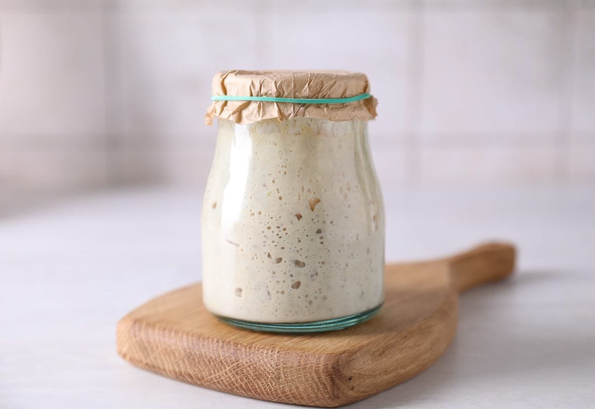 A glass jar of sourdough starter on a wooden board with a parchment paper cover.