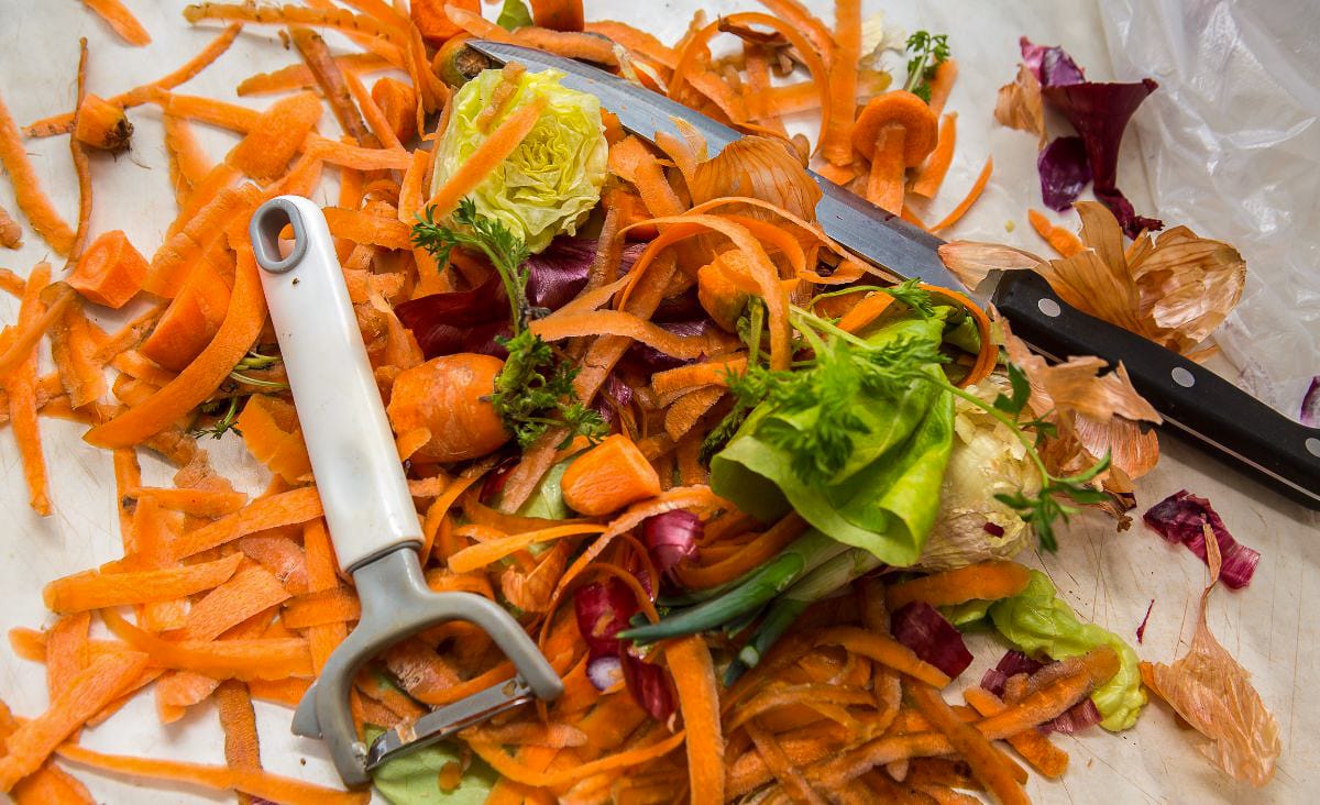 A pile of colorful vegetable scraps, including carrot peels, lettuce cores, and onion skins, with a knife and peeler on a kitchen counter.