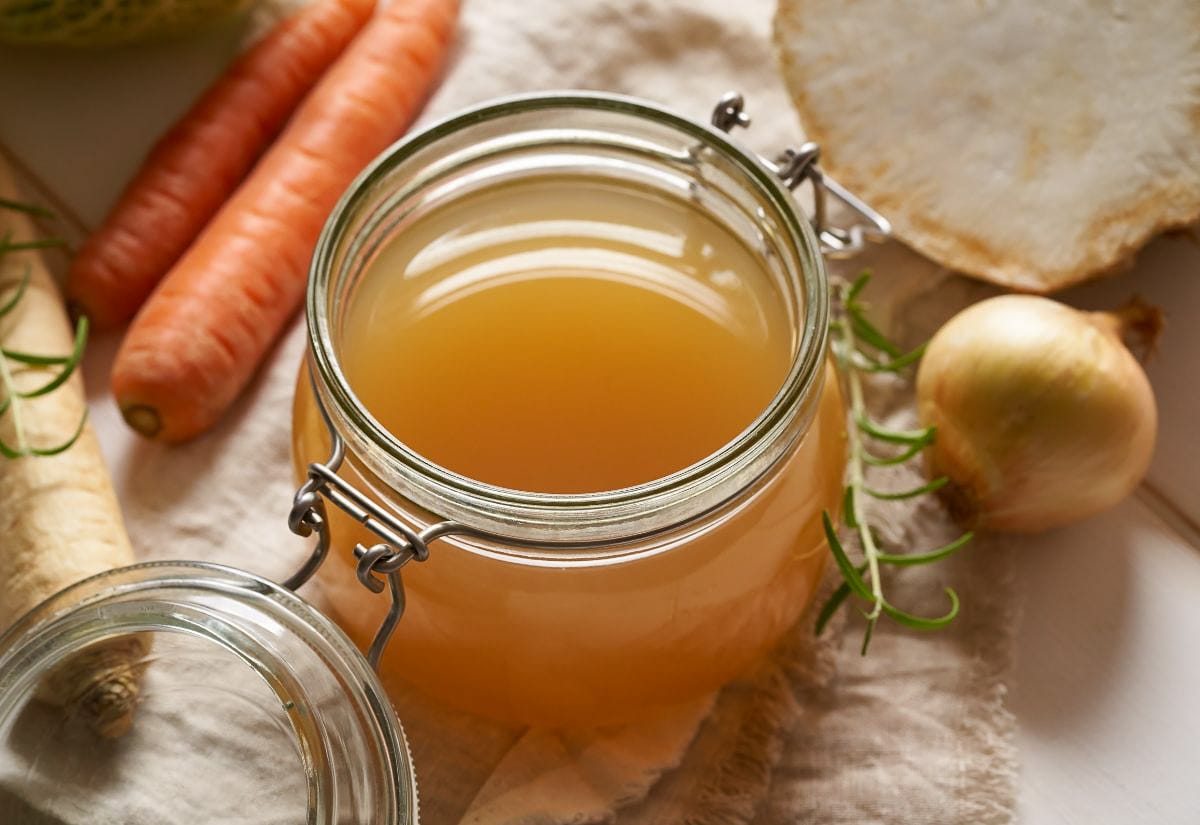 A glass jar filled with homemade vegetable broth sits next to fresh carrots, onions, and herbs.
