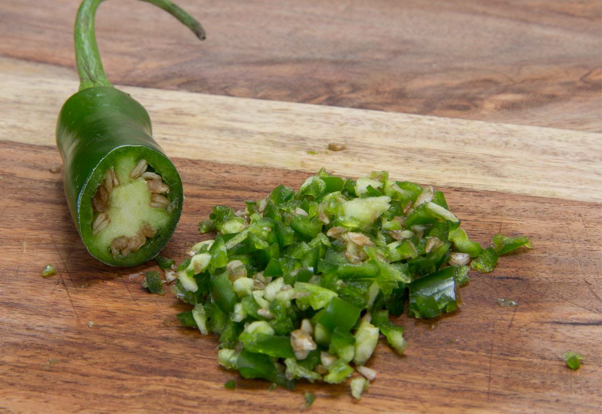 Fresh jalapeño pepper partially sliced with seeds visible, next to a pile of finely chopped jalapeño on a wooden cutting board.