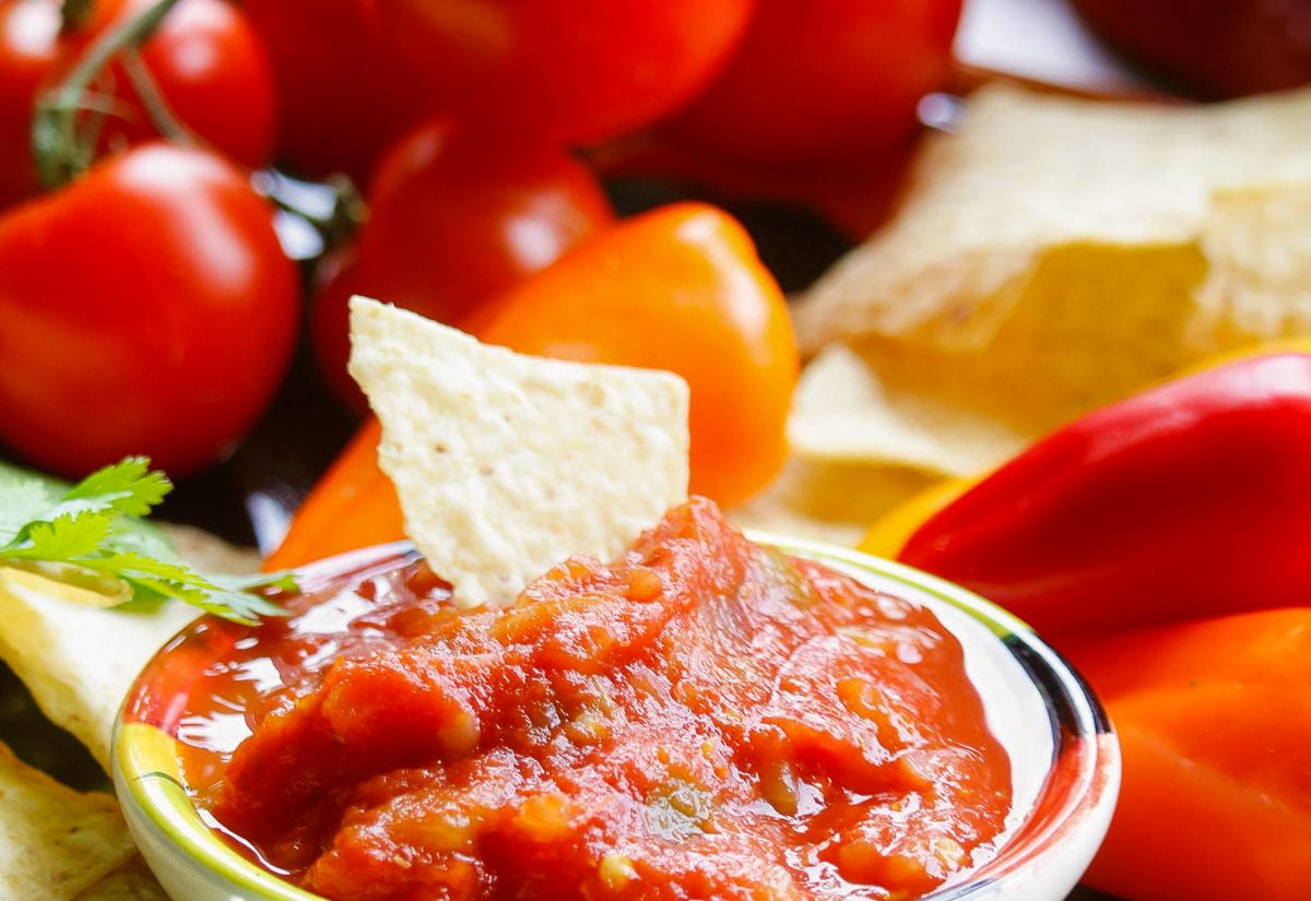 Bowl of chunky red salsa with a tortilla chip dipped in, surrounded by fresh tomatoes, mini bell peppers, cilantro, and chips in the background.